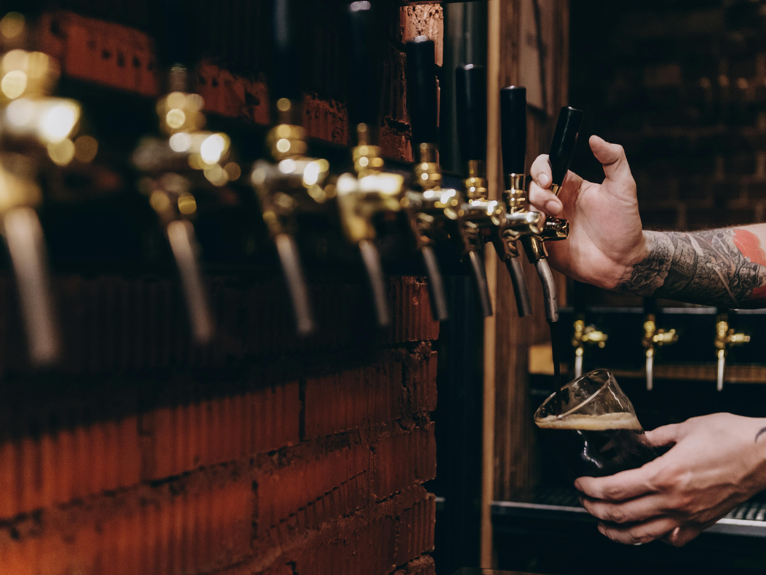 Person pouring beer from a tap at a bar, tattooed arm visible, with a glass of beer being filled.