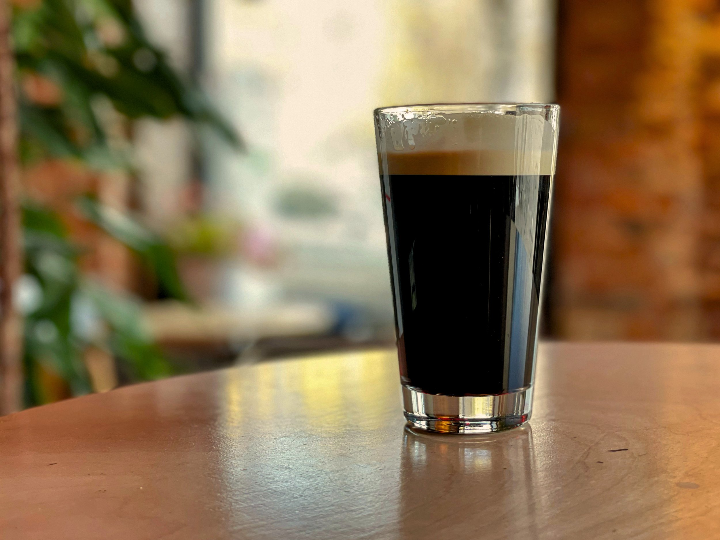 A glass of dark coffee with a frothy top sitting on a wooden table.