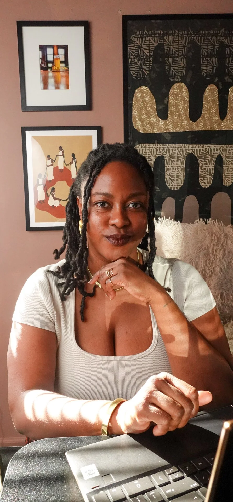 A woman with dark skin and black dreadlocks sitting at a table, smiling confidently at the camera. She is wearing a light-colored top, gold jewelry, and has dark lipstick. Behind her is a wall with framed artwork and a large piece of black, gold, and beige abstract art. There is a fluffy beige pillow on her right side.