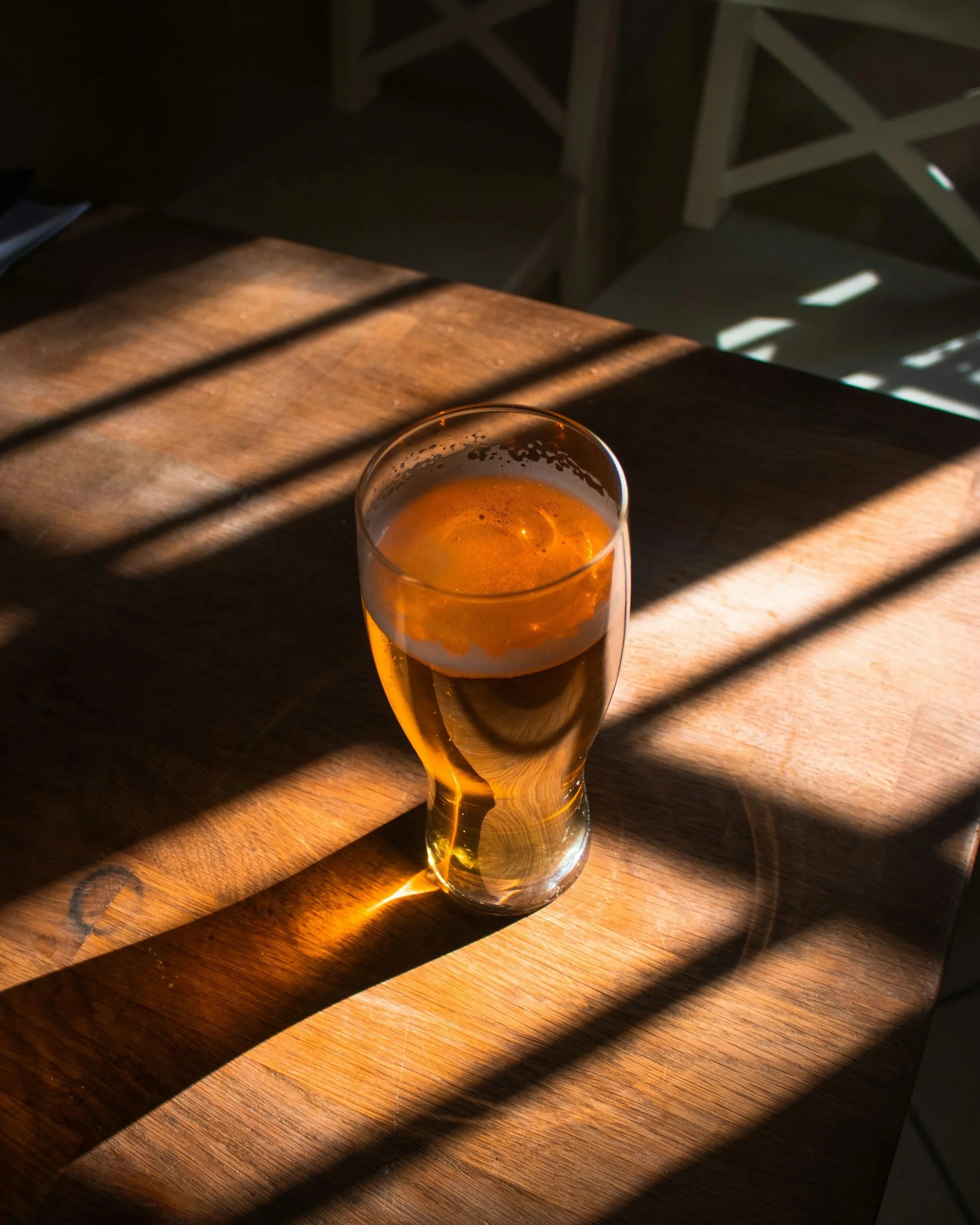 A glass of beer on a wooden table with sunlight and shadows creating patterns across the surface.