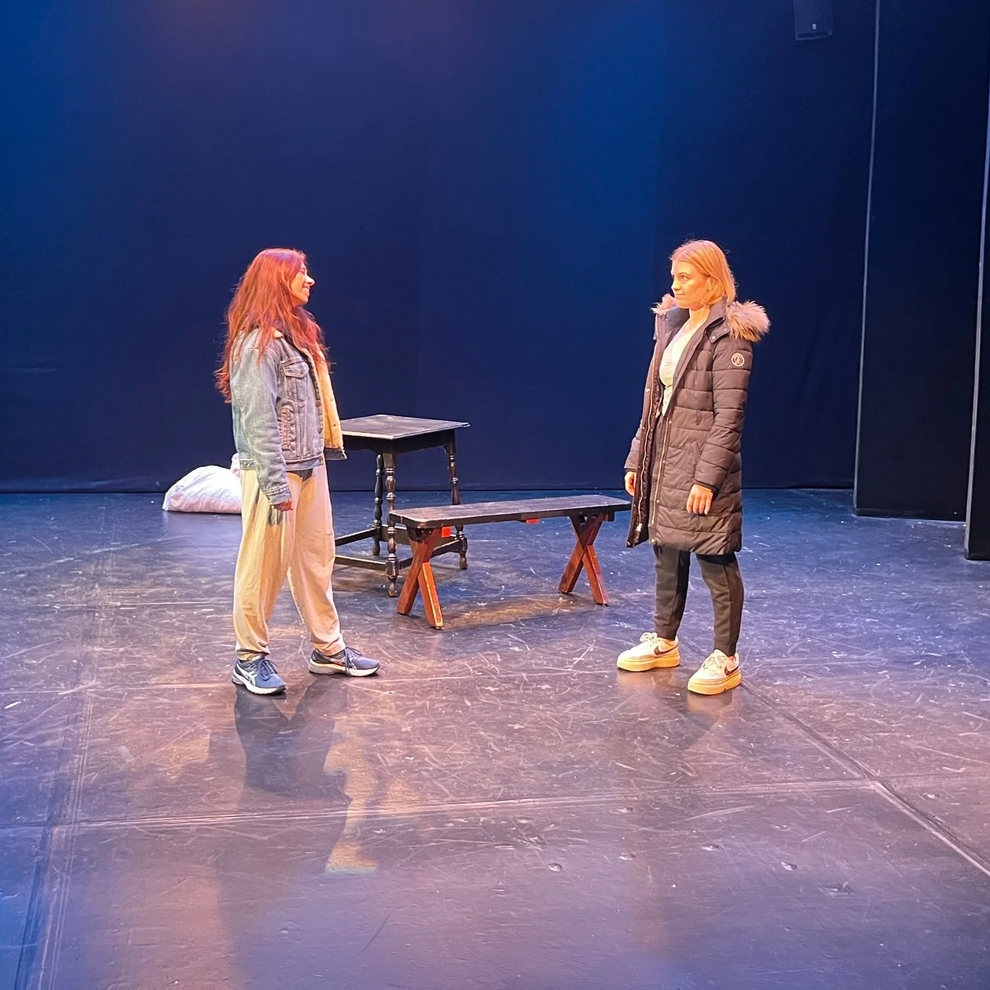 Two women with casual clothing standing on stage facing each other, with wooden tables and a dark backdrop.