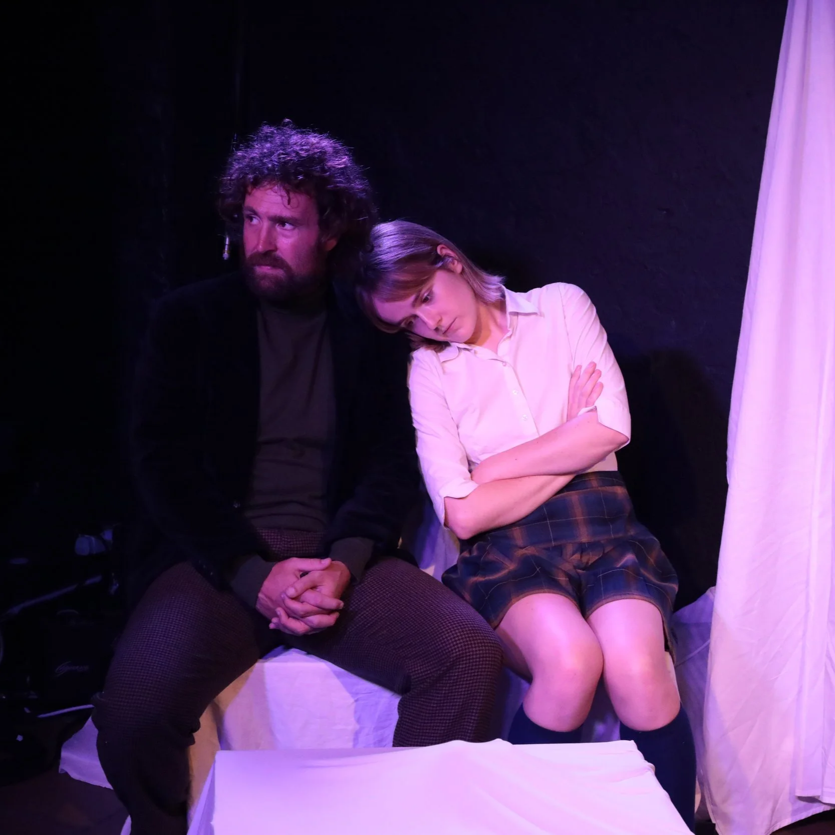 Two people sitting against a black background, one with curly hair and a beard, the other with straight hair, both looking concerned or sad, with the woman resting her head on the man's shoulder.