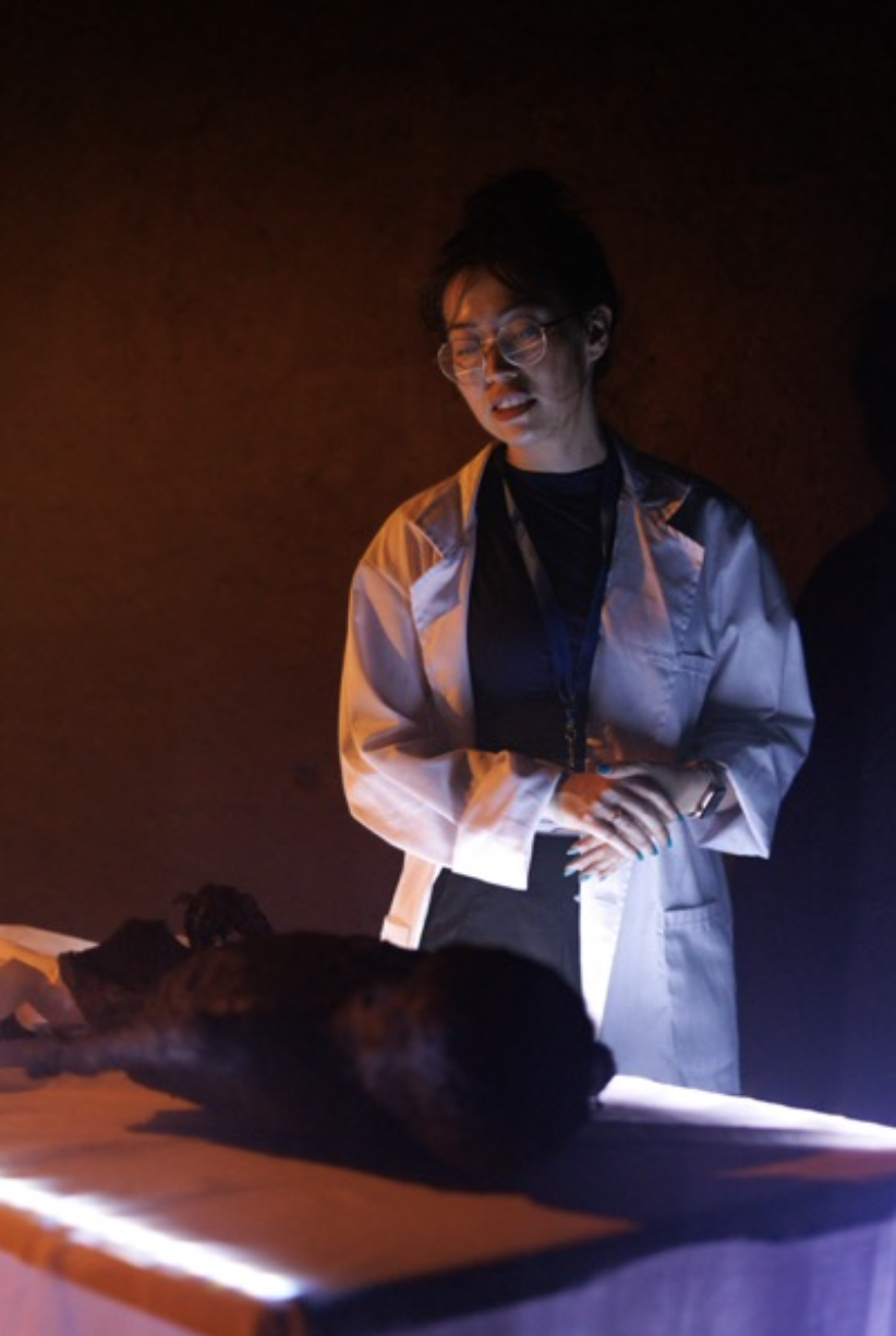 A woman in glasses wearing a light-colored lab coat looks at a large object on a table in a dark room with dramatic lighting.
