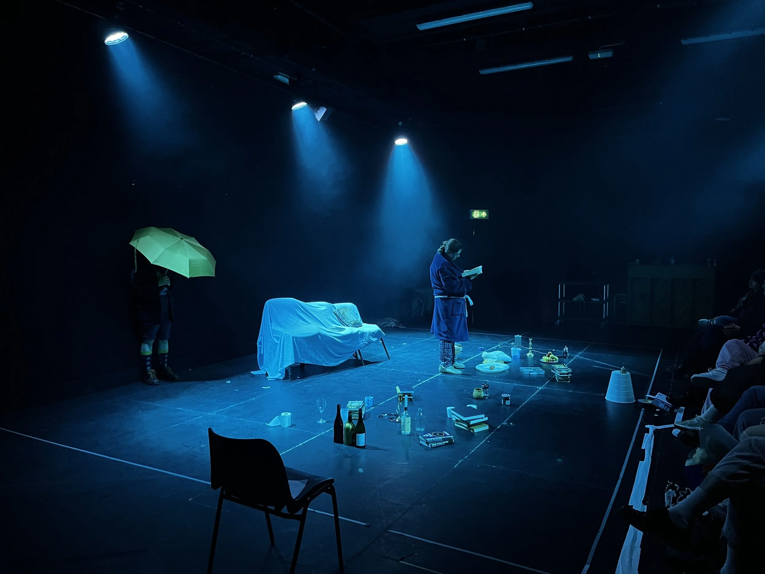A theatrical stage featuring a person reading a book, various scattered props including bottles, books, and a covered couch, and a small audience seated on the right side, with dramatic blue lighting.