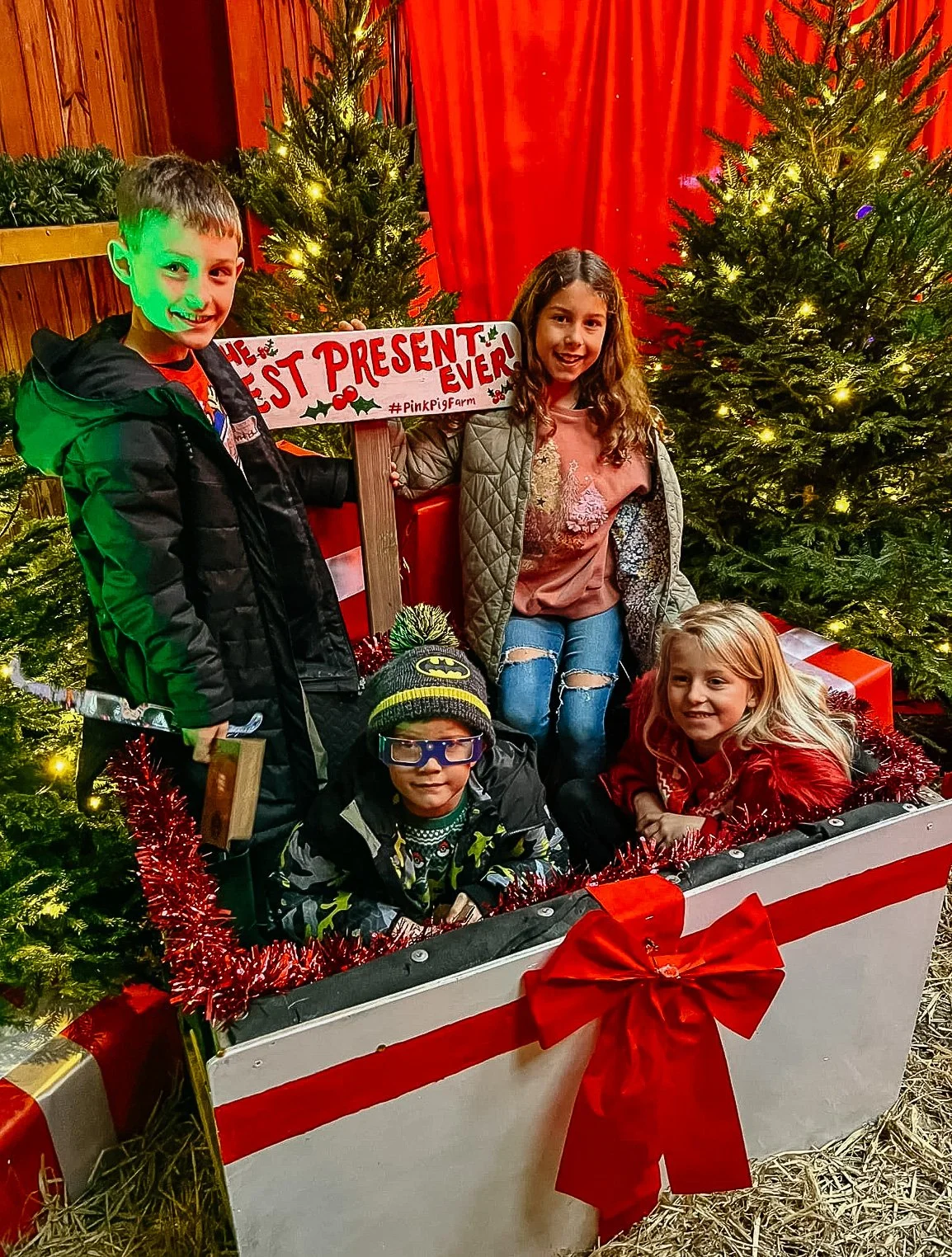 Children posing in a Christmas-themed setting with decorated Christmas trees and a sign that reads 'Best Present Ever!'