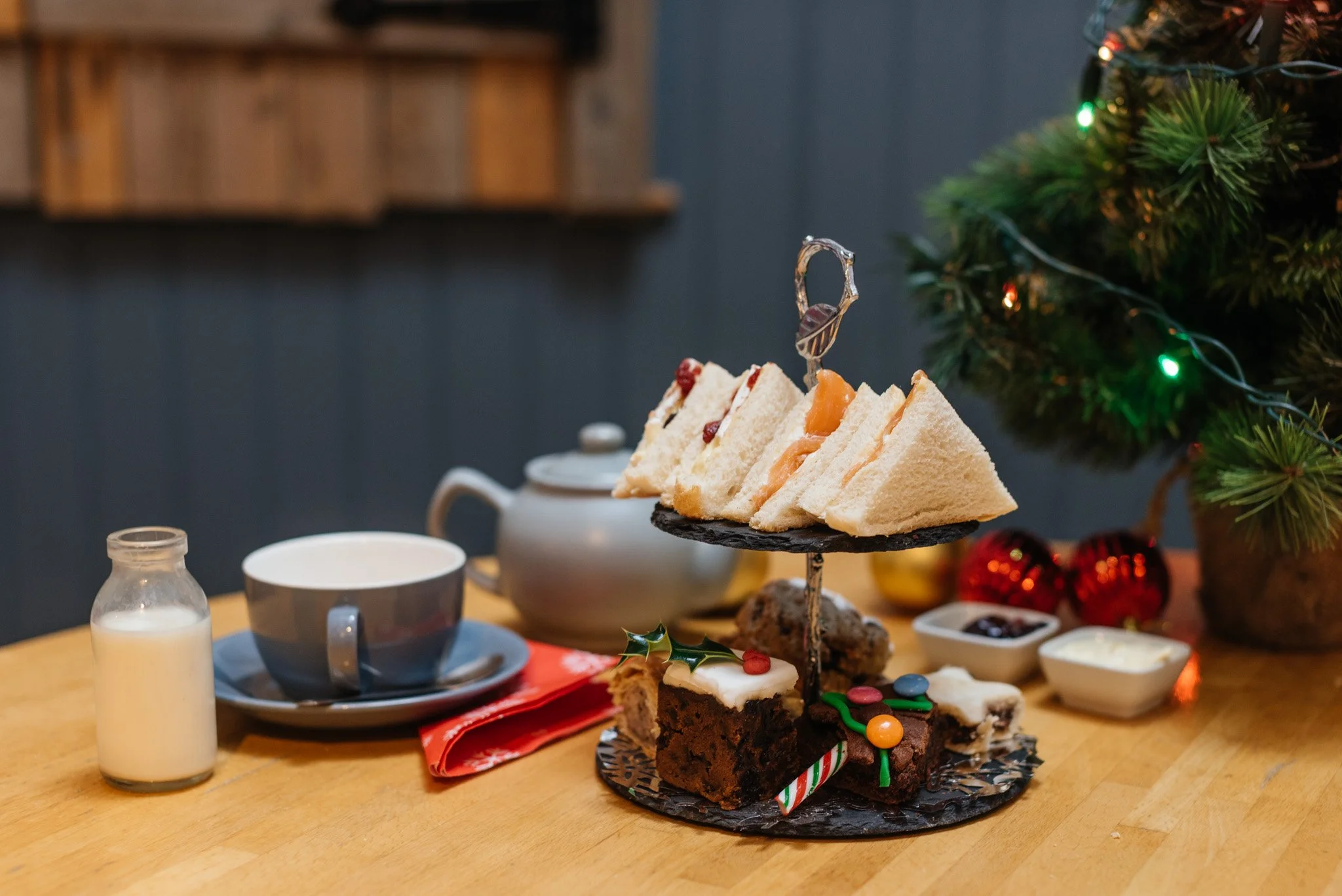 A tiered tray of assorted holiday cookies and treats on a wooden table, with a Christmas tree decorated with lights in the background.