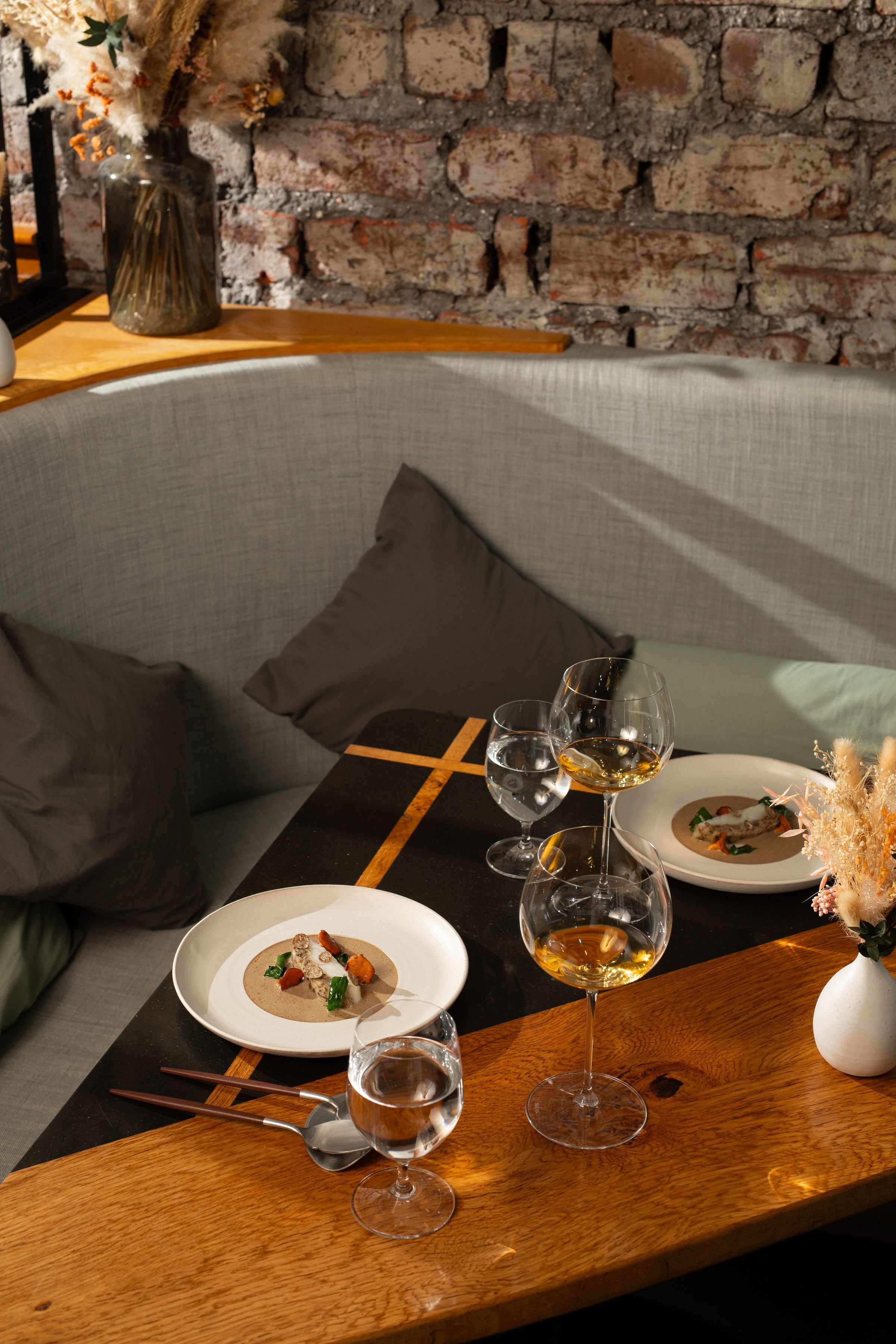Elegant restaurant table with plated dishes, wine glasses, silverware, dried floral arrangements, and a brick wall backdrop.