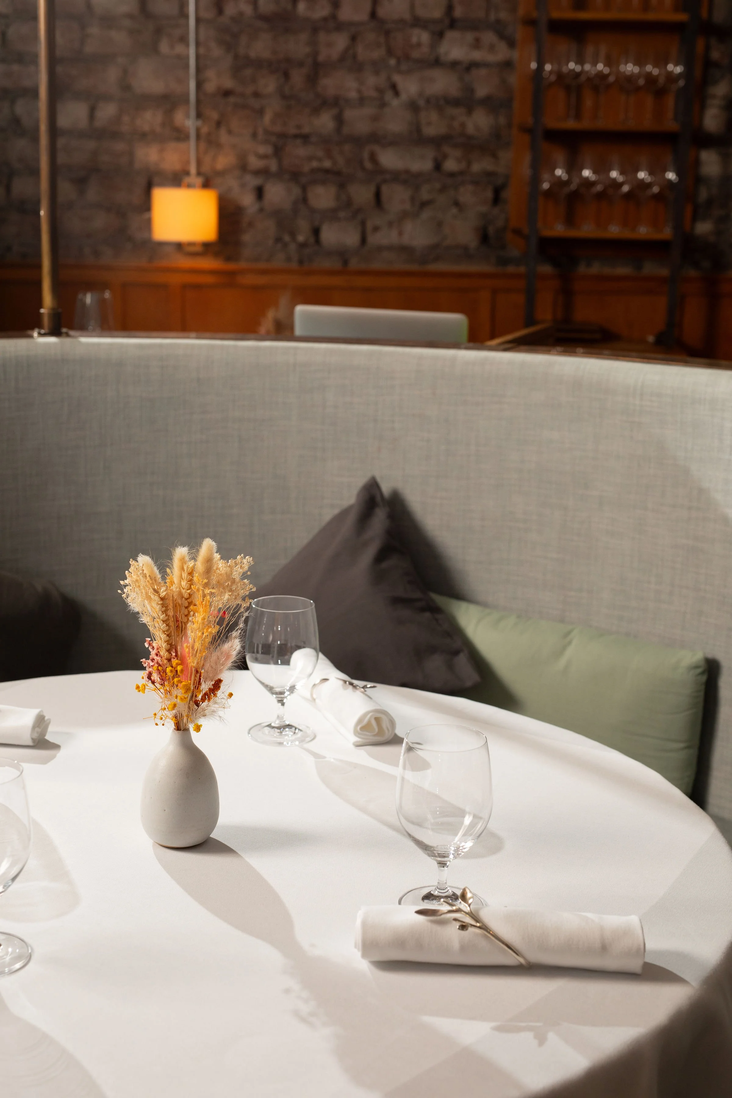 Elegant restaurant dining table with white linens, wine glasses, rolled napkins, dried flowers, and a warm brick-and-wood interior