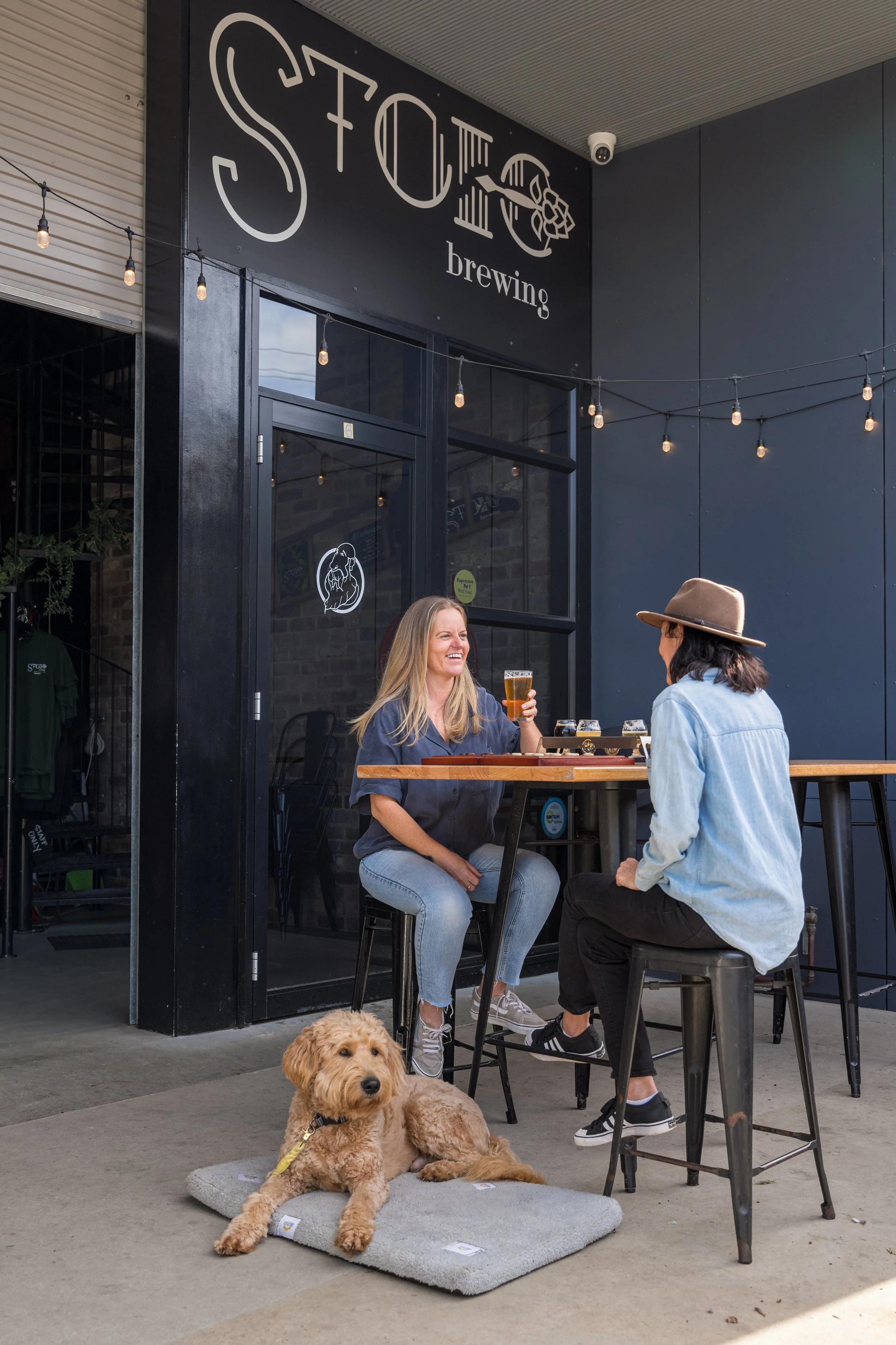 Two women sitting outdoors at a bar table, engaging in conversation. One woman is holding a glass of beer and smiling, wearing a gray shirt and light jeans. The other woman has dark hair, wears a light blue jacket, black pants, and a wide-brimmed hat. A golden retriever is lying on a gray mat on the ground in front of them, looking toward the camera. The scene is outside a brewery named 'STOIC brewing'.