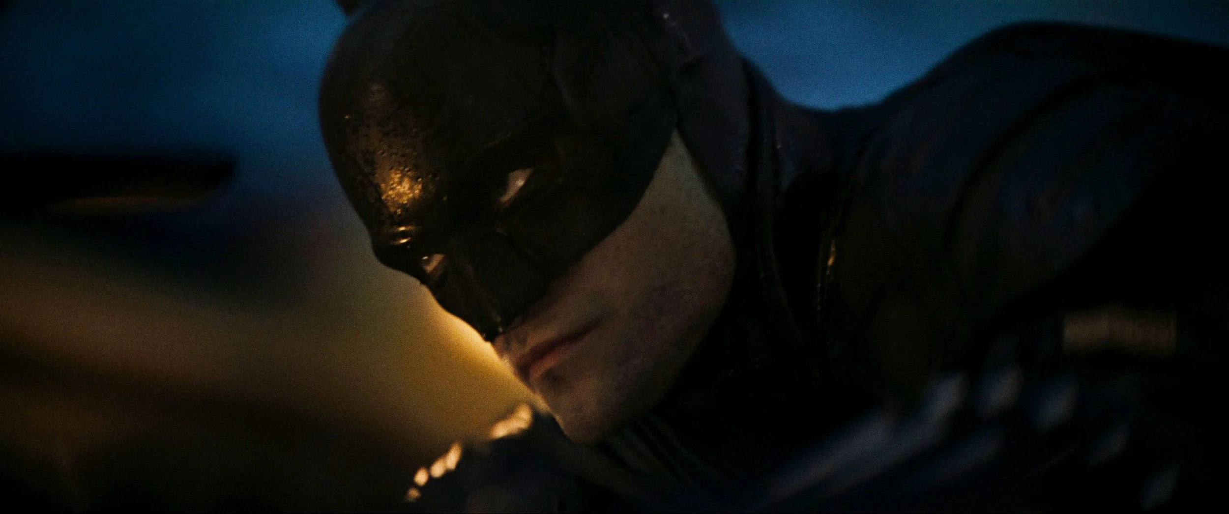 Close-up of a person wearing a black mask and dark clothing, with a dark background, possibly in a dimly lit environment.