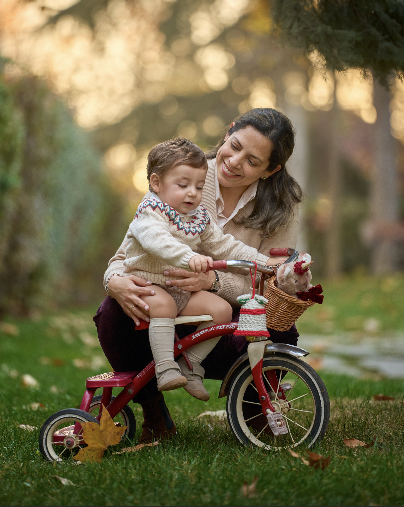 Una mujer y un niño pequeño en un parque, montados en una bicicleta con ruedas de entrenamiento, disfrutando de un día de otoño con hojas caídas.