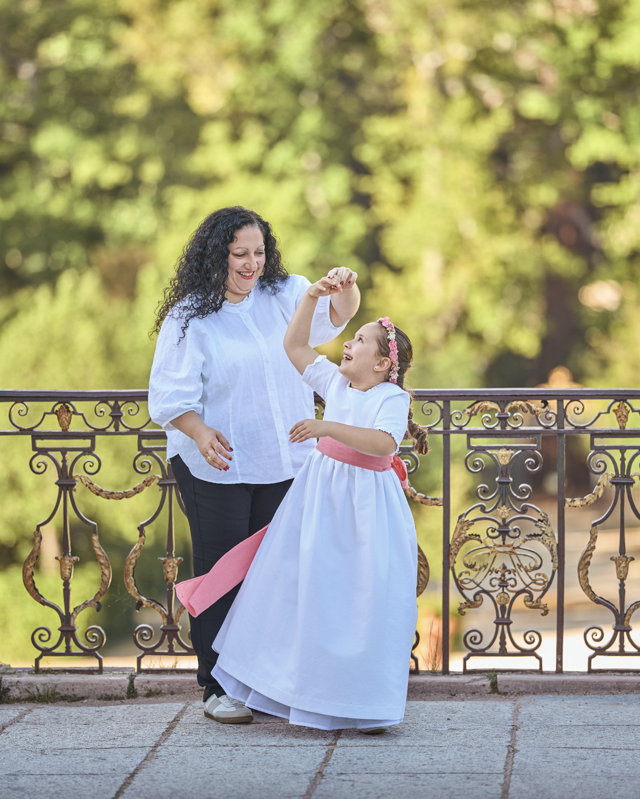 Una mujer adulta y una niña bailando juntas en un parque, la niña lleva un vestido largo blanco con un lazo rosa y una corona de flores, la mujer lleva blusa blanca y pantalones negros, riendo y disfrutando del momento, con fondo de árboles verdes y 