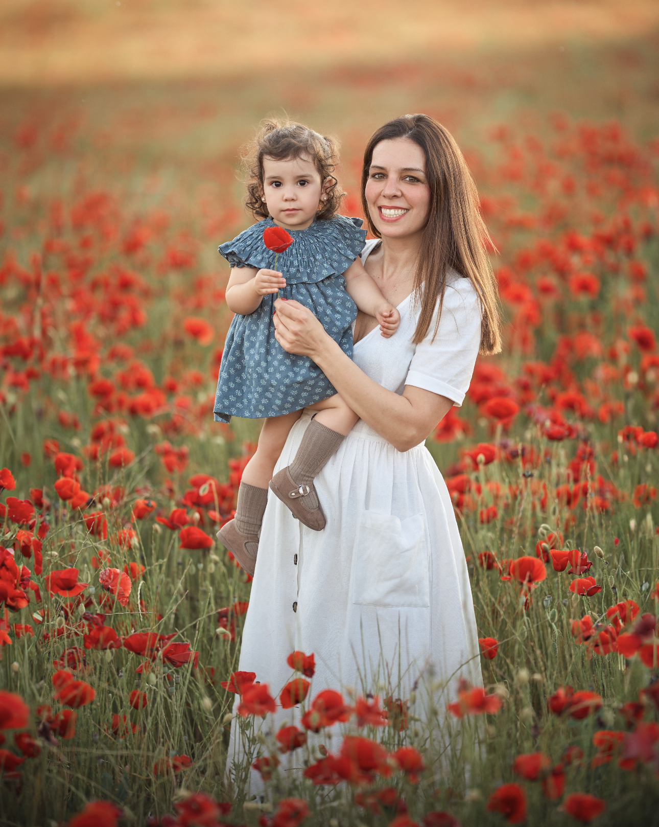 Mujer joven sosteniendo a una niña en un campo de amapolas rojas durante el atardecer