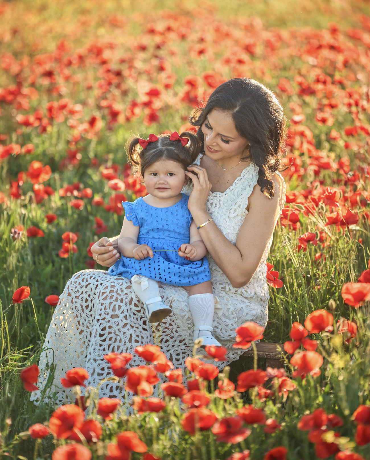 Mujer y niña en un campo de amapolas durante la tarde, la mujer viste un vestido blanco de encaje y la niña lleva un vestido azul. La mujer sostiene a la niña en su regazo y ambas están sonriendo, disfrutando del momento en un paisaje natural lleno d
