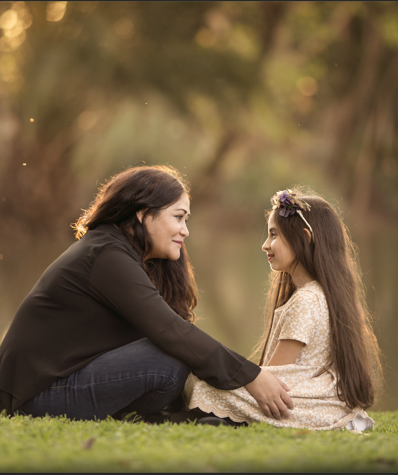 Mujer y niña sentadas en el césped, mirándose y sonriendo con fondo de árboles y luz dorada del atardecer.