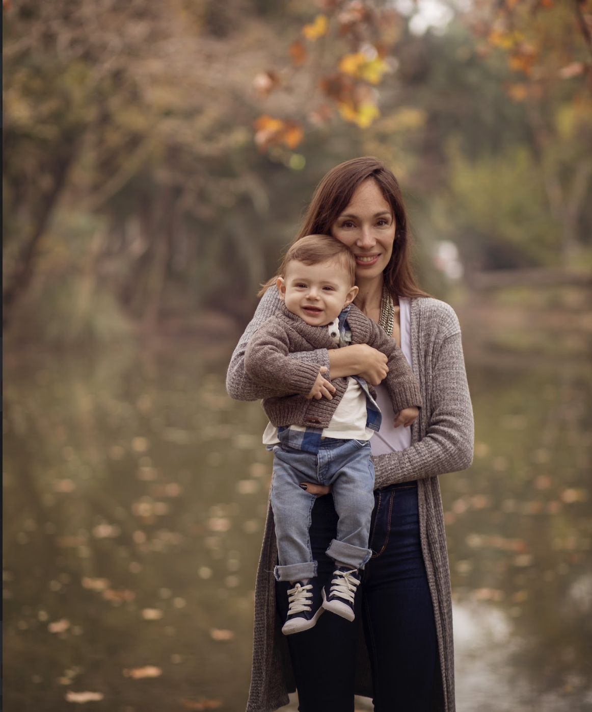 Una mujer y un niño pequeño en un entorno natural con árboles y agua, en otoño.