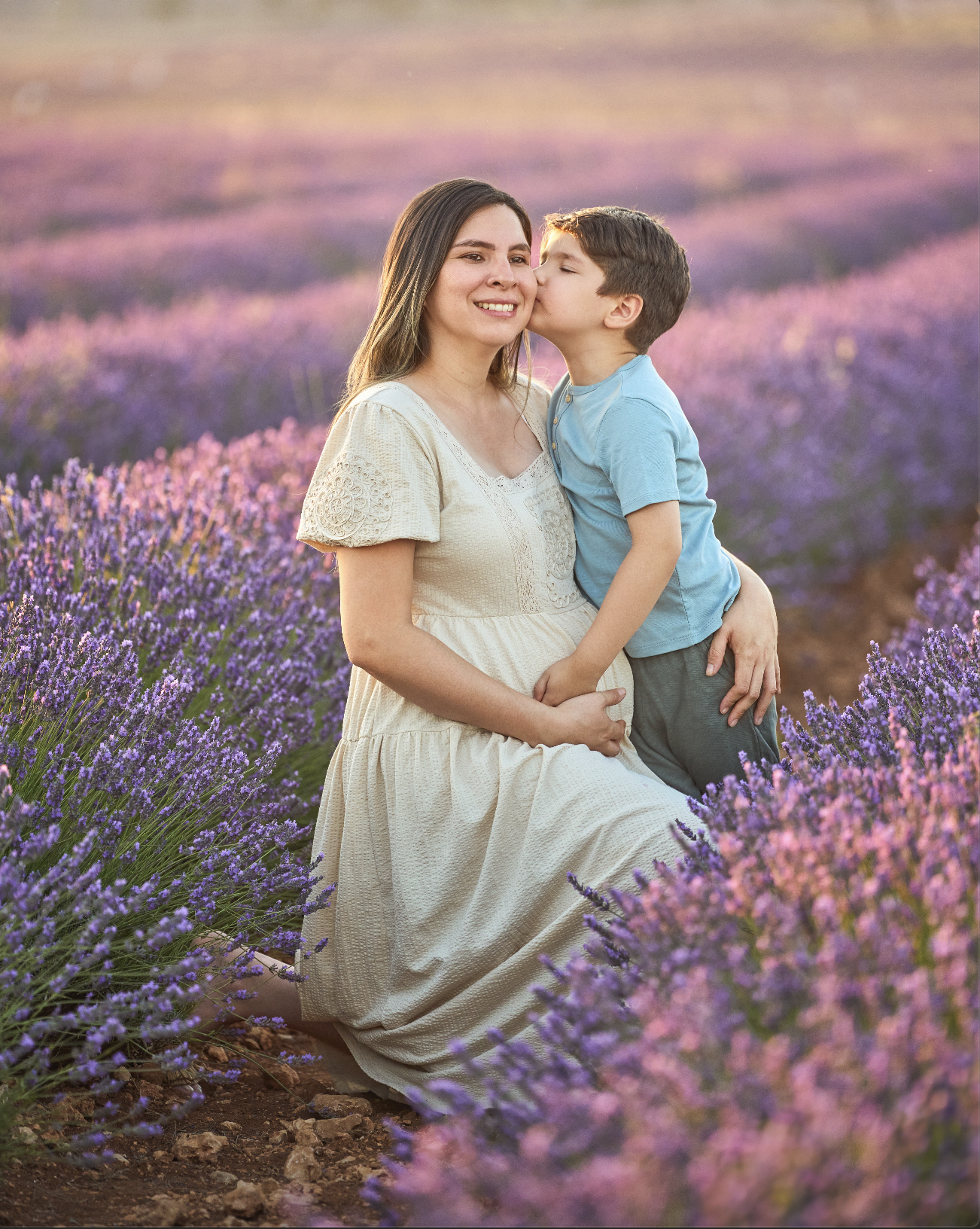 Mujer y niño en un campo de lavanda, la mujer está sonriendo mientras el niño le da un beso en la mejilla, al atardecer.