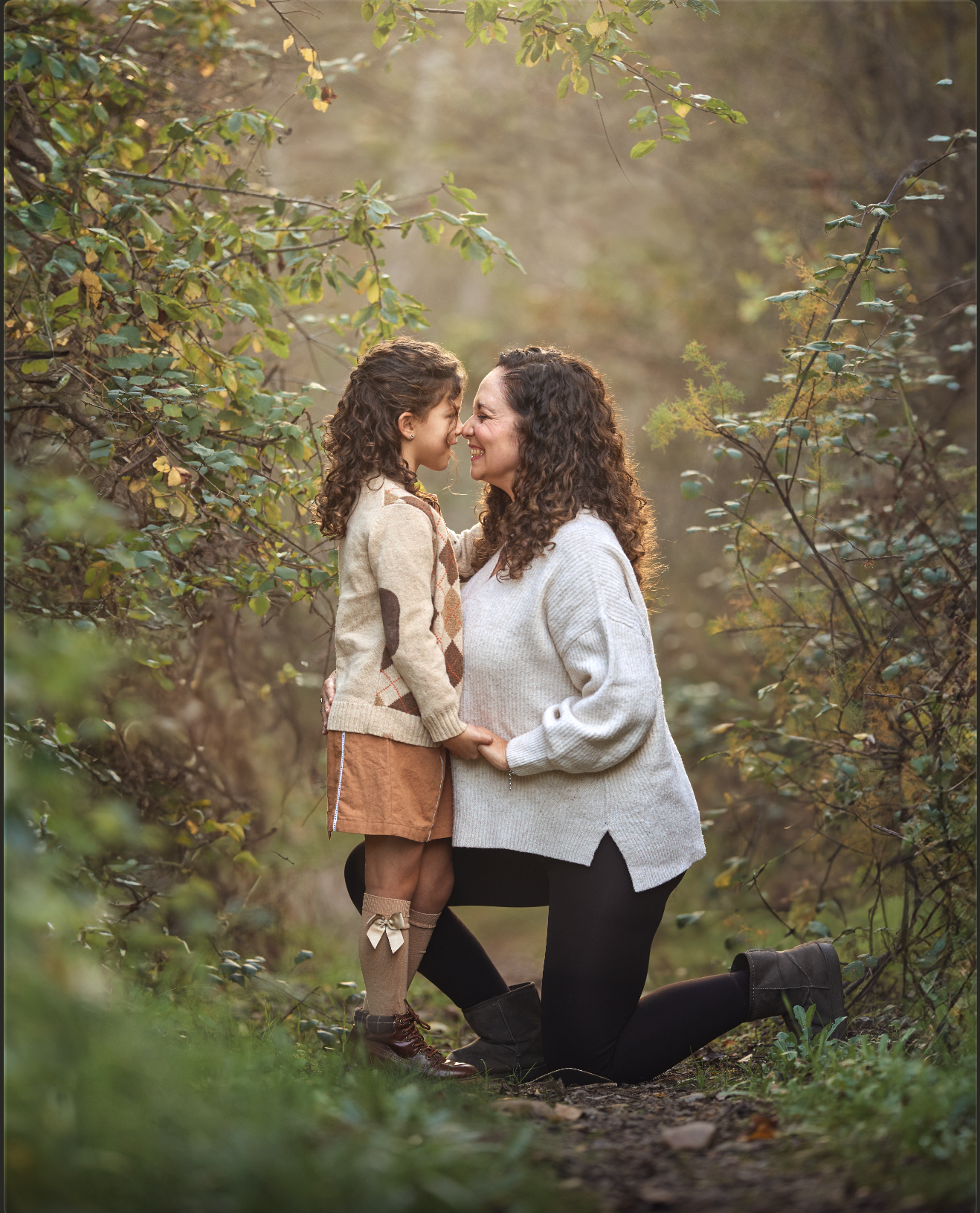 Una mujer y una niña se abrazan y se tocan las narices en un bosque con árboles y hojas verdes y doradas, disfrutando un momento juntos al atardecer.