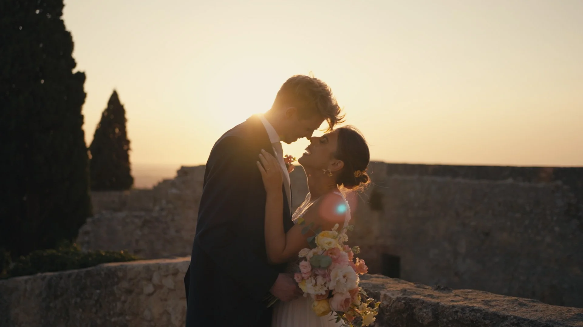 A couple in wedding attire sharing an intimate moment at sunset, with the bride holding a bouquet of flowers.
