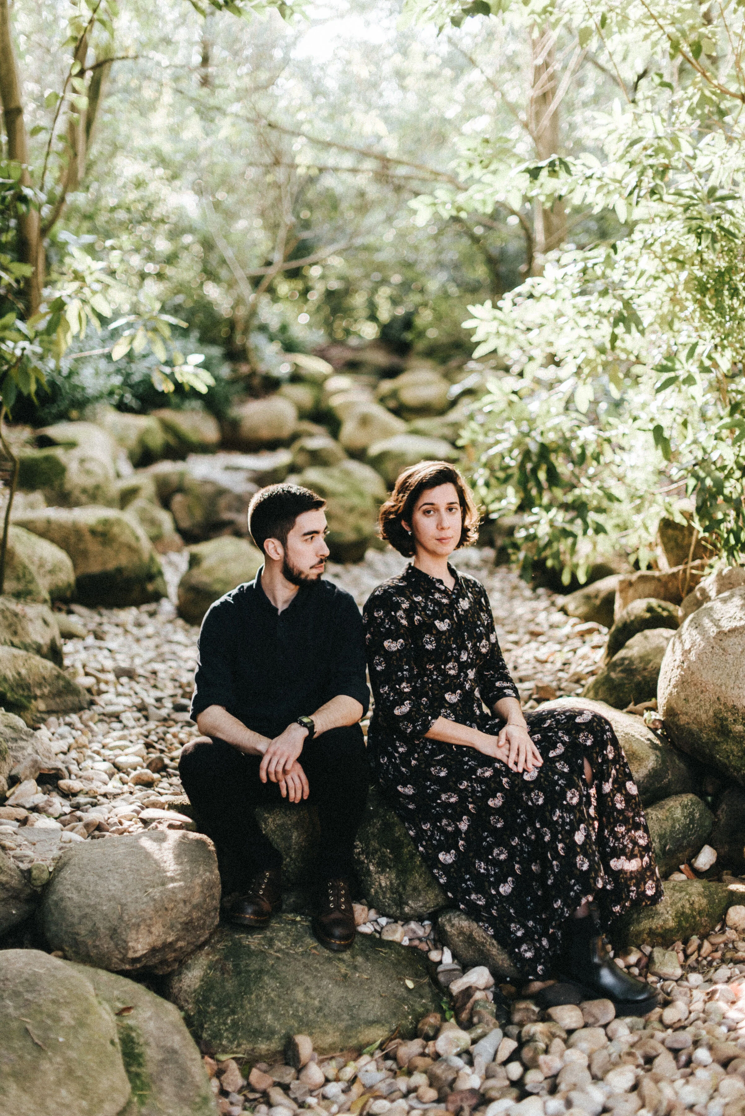 A man and woman sitting on rocks in a lush, sunlit forest with a rocky creek bed surrounding them.