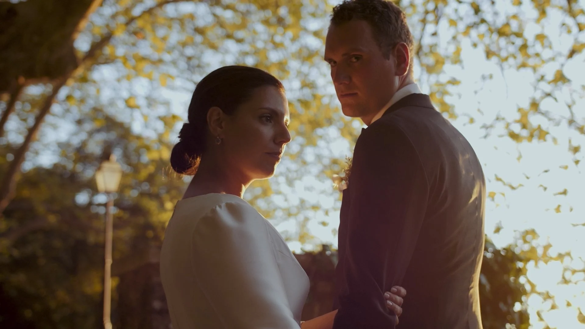 A man and woman dressed formally, standing close together outdoors during sunset, looking serious.