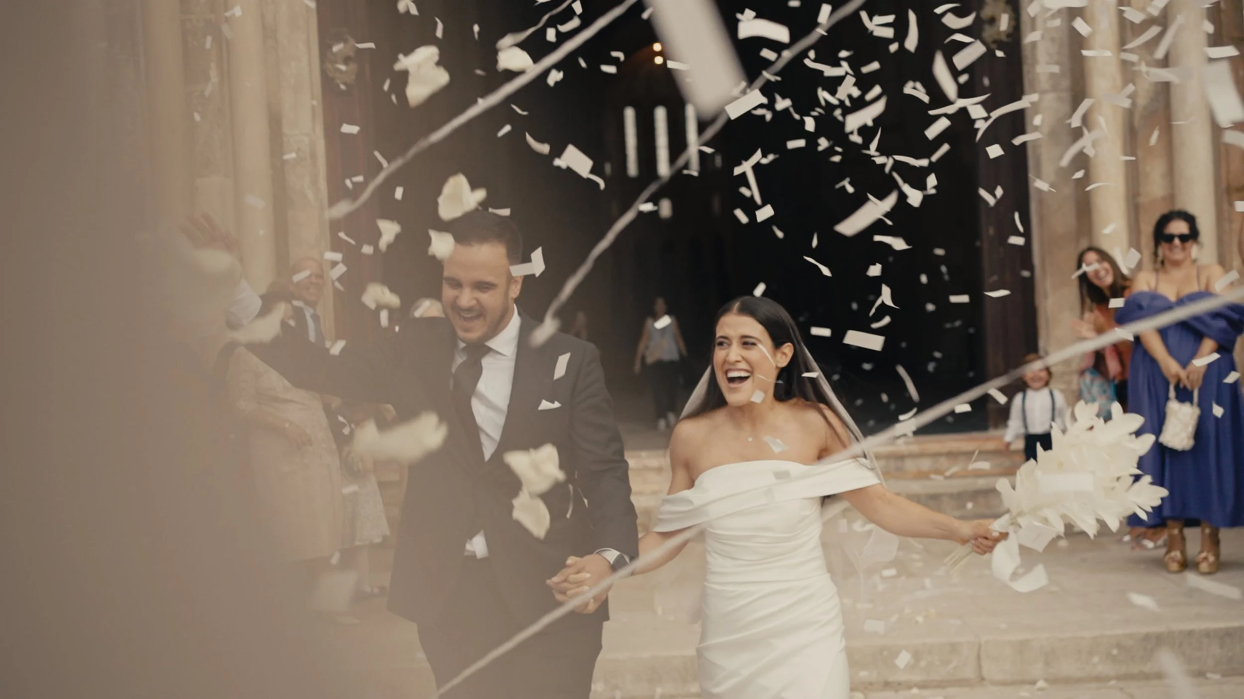 A newlywed couple smiles and runs hand in hand, celebrating their wedding with guests and confetti outside a church.