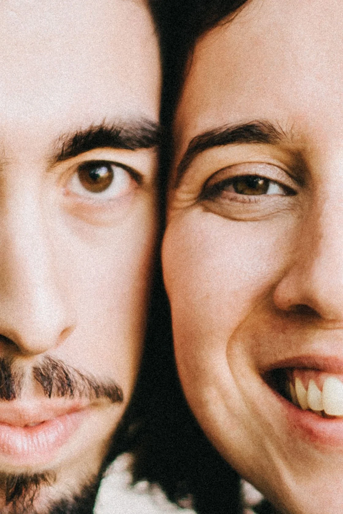 Close-up of two people's faces, one male and one female, positioned tightly together, both smiling and showing their teeth.