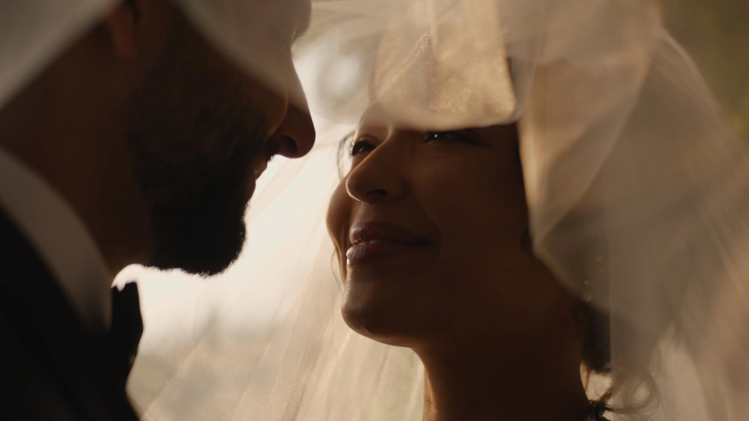 A close-up of a man and woman leaning close to each other, smiling, with a soft light illuminating their faces, behind a translucent fabric or curtain.