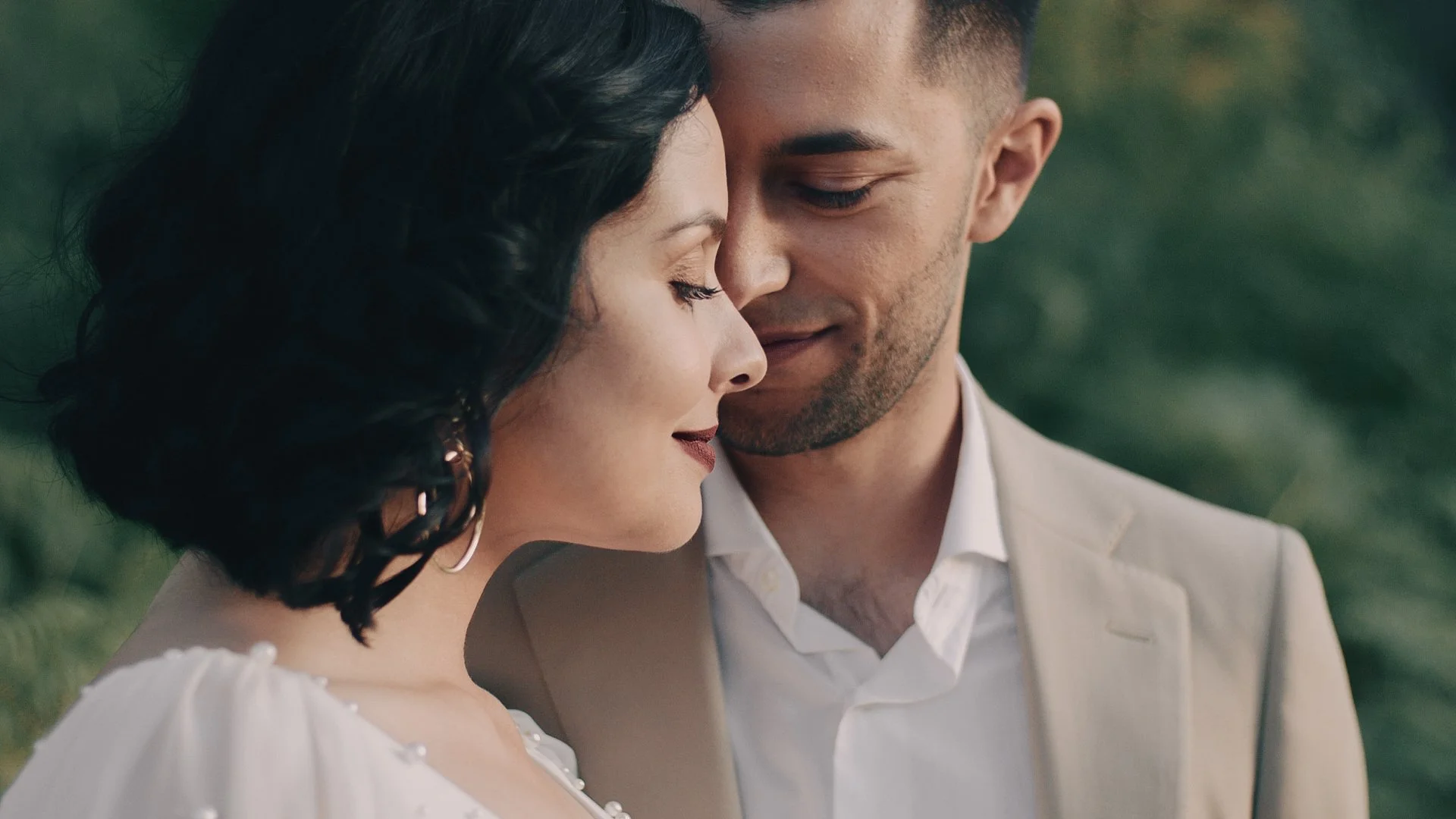 A close-up of a man and woman with foreheads touching, eyes closed, smiling softly outdoors.