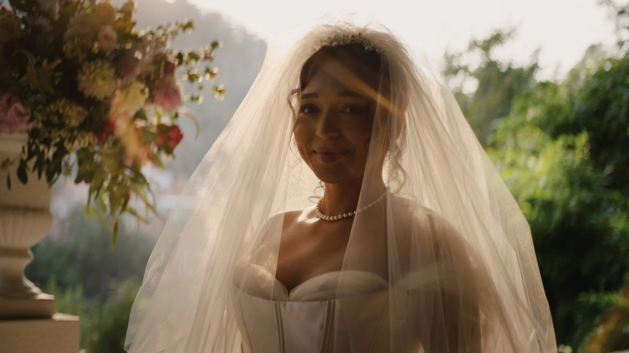 A bride in a wedding dress and pearl necklace, smiling softly, with a veil over her face, standing outdoors near a large flower arrangement with trees and sunlight in the background.