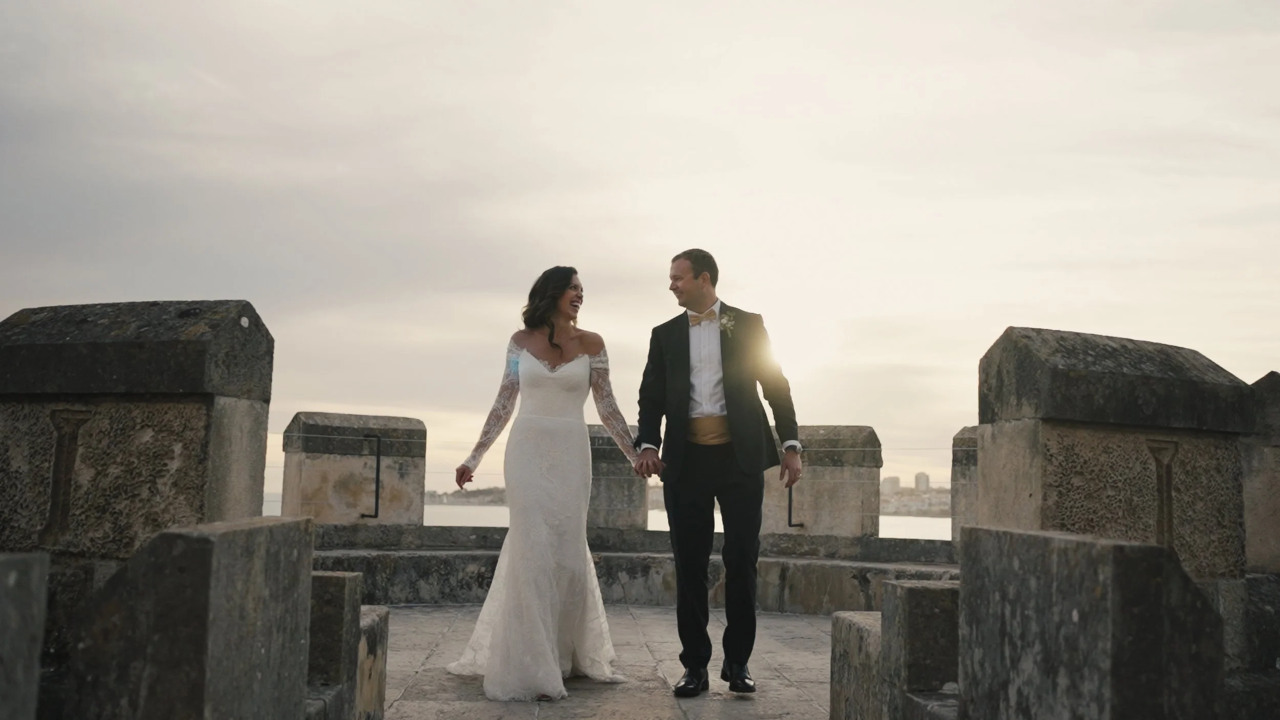 Bride and groom walking hand in hand on a stone platform outdoors during sunset or sunrise, with a city skyline in the background.
