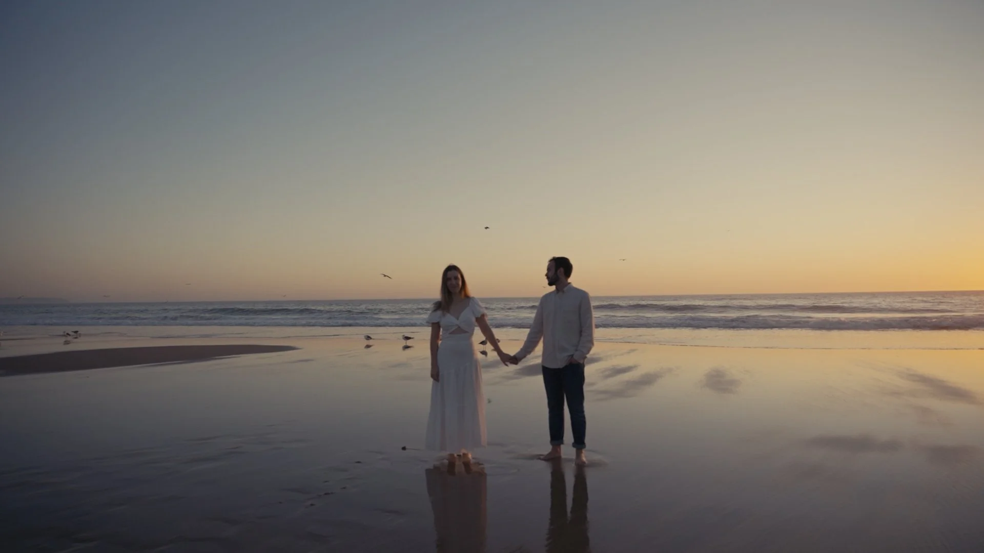A couple holding hands on the beach at sunset, with seagulls flying overhead and their reflections visible in the wet sand.