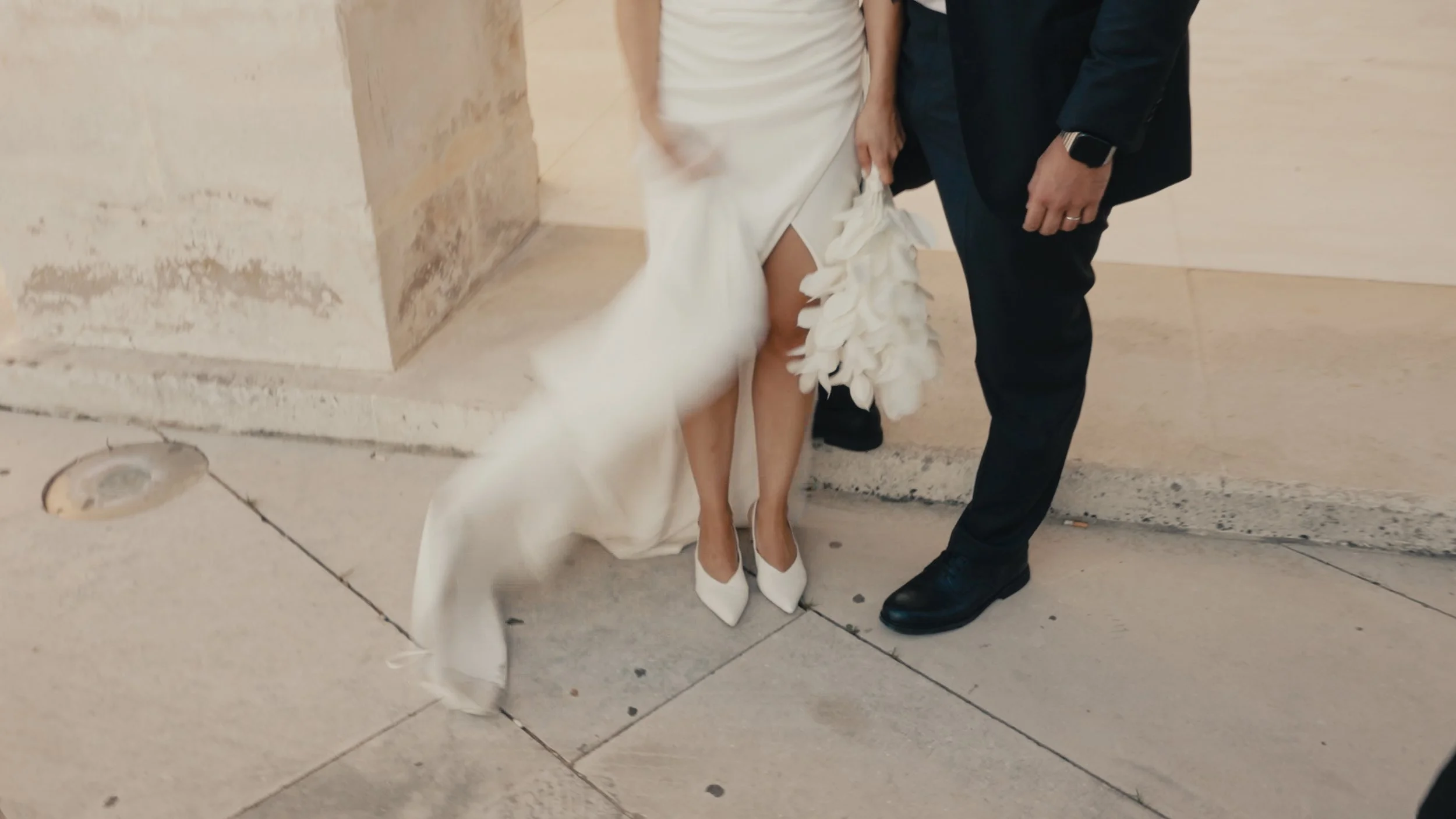 Close-up of a bride and groom's lower bodies, standing on a sidewalk, with the bride wearing a white wedding dress and high heels, and the groom in black shoes and trousers, holding a floral accessory.