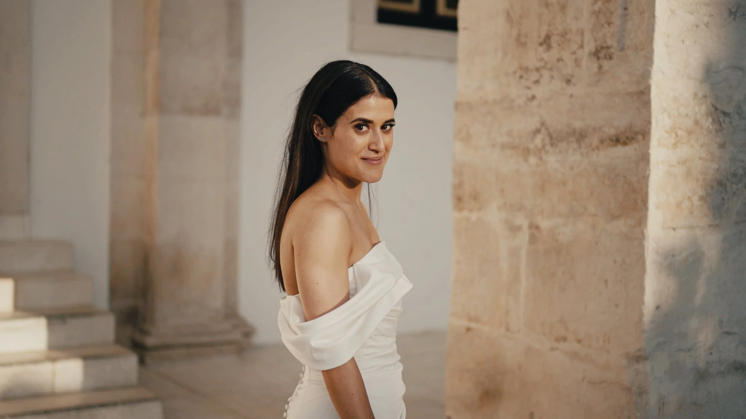 A woman with long dark hair wearing a white off-the-shoulder dress standing in an indoor space with stone walls and stairs.
