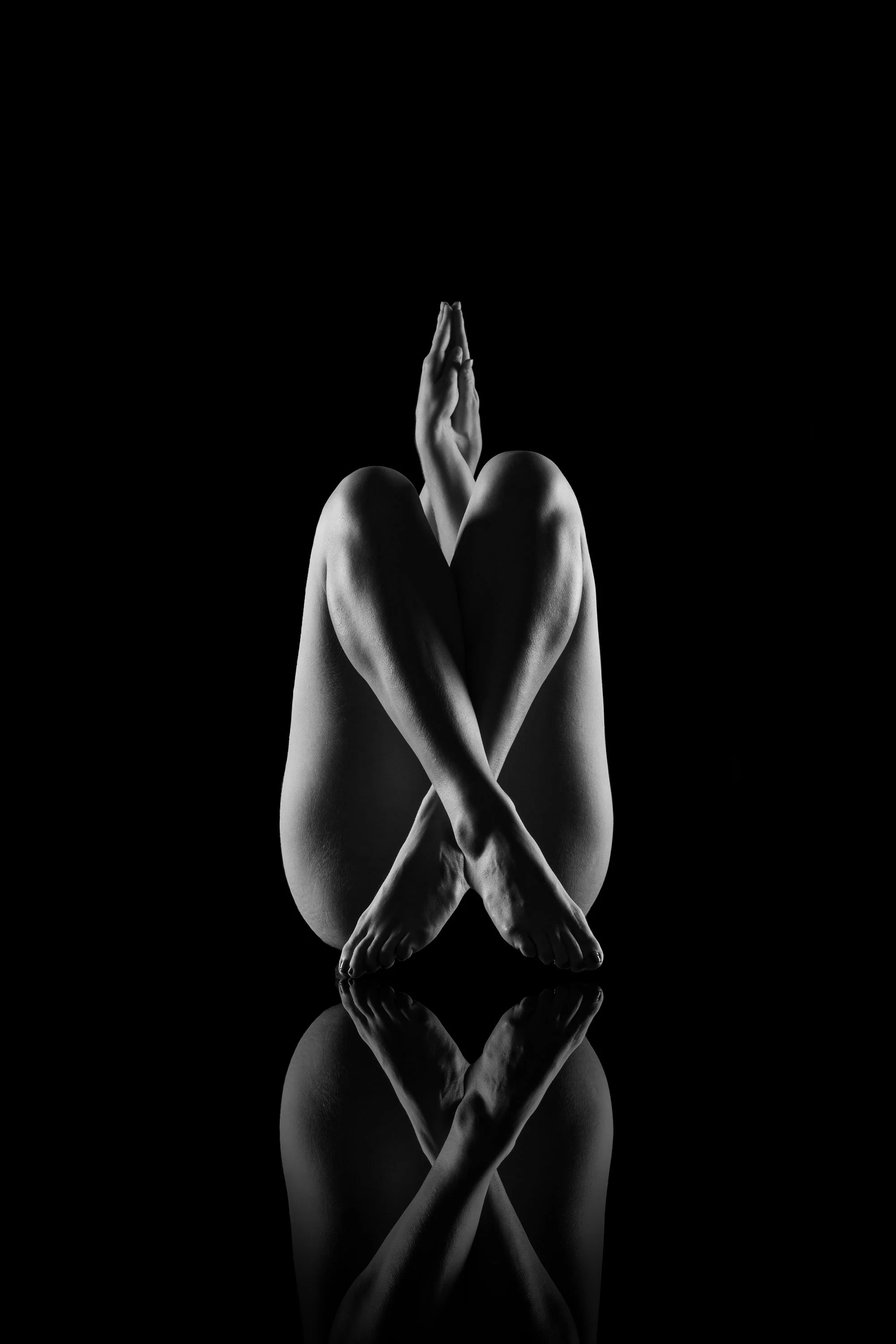 Schwarz-weiß-Fotografie eines verschlungenen Körpers auf spiegelnder Oberfläche, vor dunklem Hintergrund.
