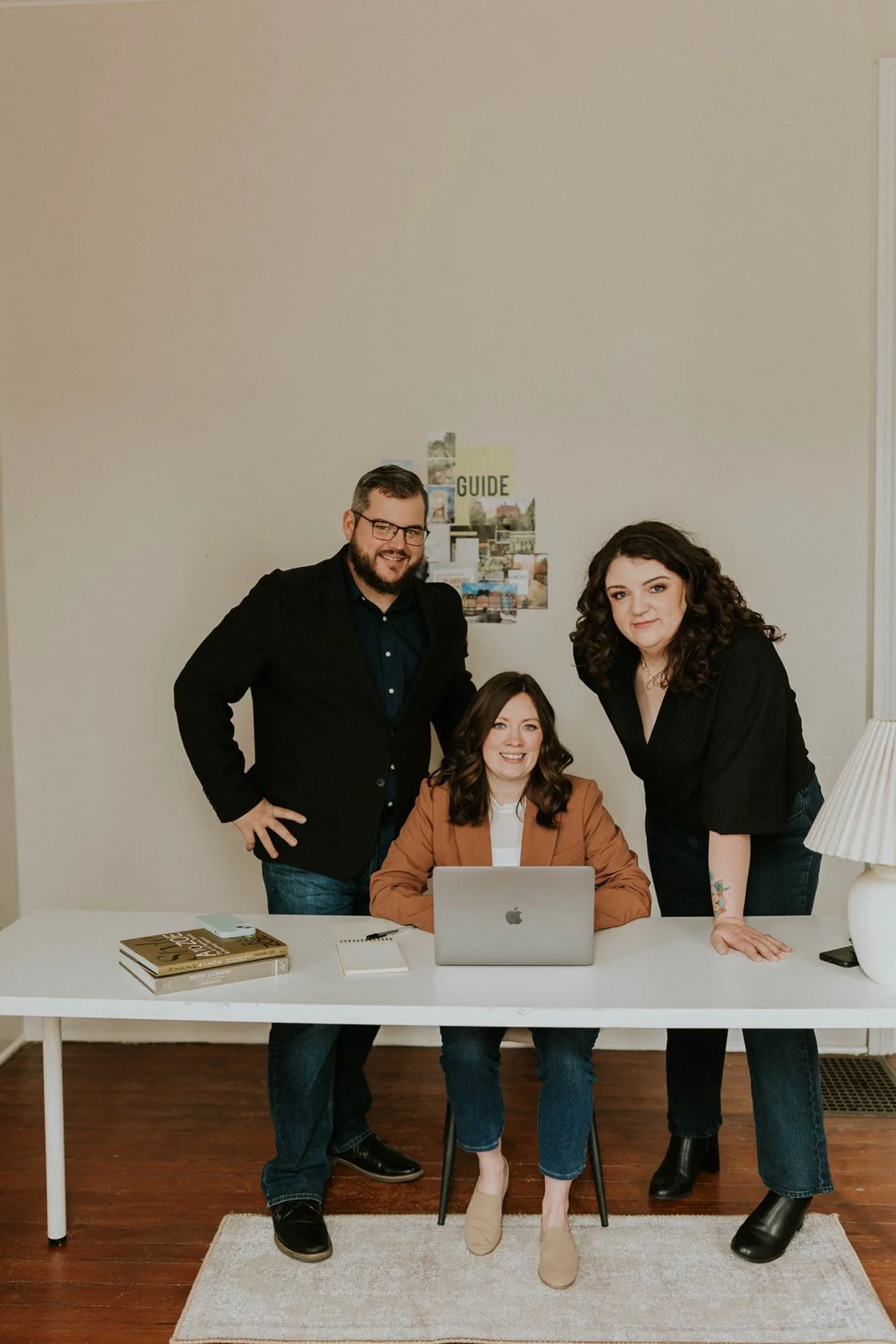 Three people, two women and one man, posing around a white desk with a laptop, books, and notebooks, in a room with beige walls and hardwood floors.