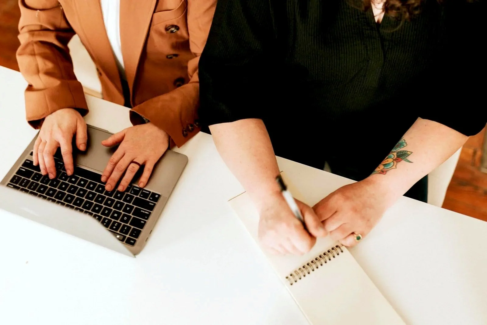 Two women are working at a white table with a laptop and a spiral notebook, one is typing on the laptop, and the other is writing in the notebook. The best KY TCs.