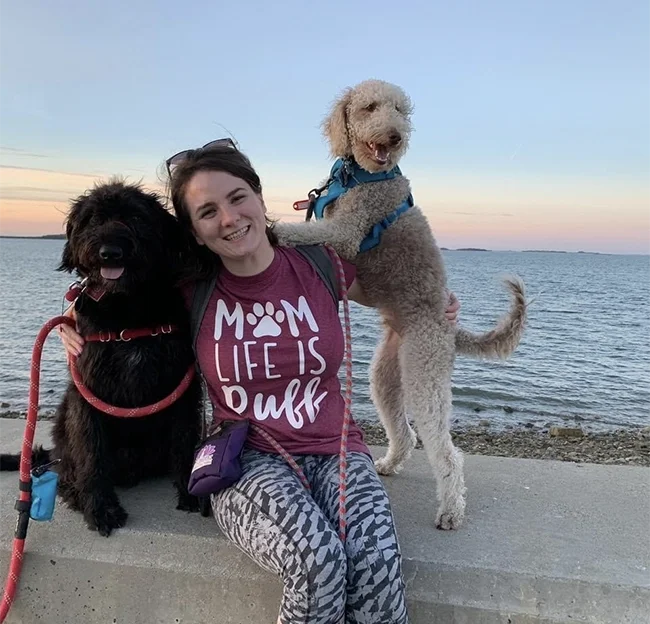 A woman with two large dogs sitting by the water at sunset, smiling. The woman wears a maroon shirt that says 'MOM LIFE IS PAWFF' and patterned leggings. One dog is black with a red leash, and the other is light-colored with a blue harness, standing on its hind legs.
