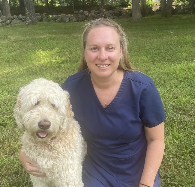 A woman in navy scrubs smiling and sitting on grass outdoors with a large, curly-haired white dog.