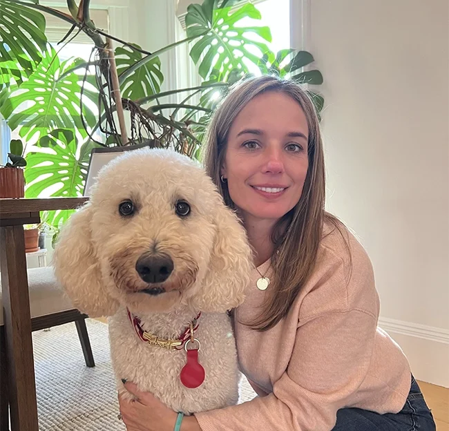 A woman with long brown hair and a light pink sweater smiling while holding a cream-colored poodle with curly fur, large dark eyes, and a red collar, in a room with big green houseplants in the background.