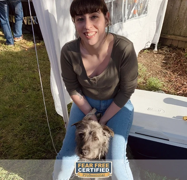 A woman with short black hair and bangs, smiling, sitting on a blue chair outdoors, holding a small gray and white dog on her lap, with a white tent and other people in the background.