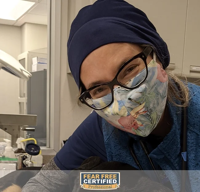 A healthcare professional wearing a blue scrubs, a dark blue head covering, glasses, and a colorful tropical face mask, inside a clinical setting.