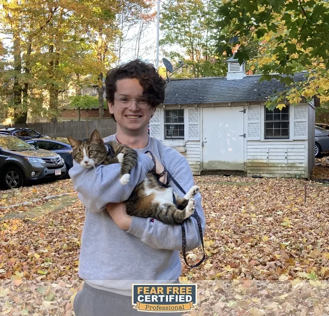 A person with short curly hair, glasses, and a gray sweatshirt smiling while holding a large tabby cat outdoors in front of a small white shed with trees and cars in the background during autumn.