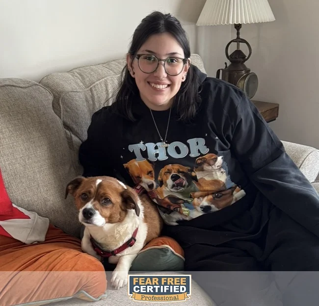Young woman with dark hair, glasses, and a Thor dog shirt sitting on a beige couch with a brown and white dog, posing for the camera.