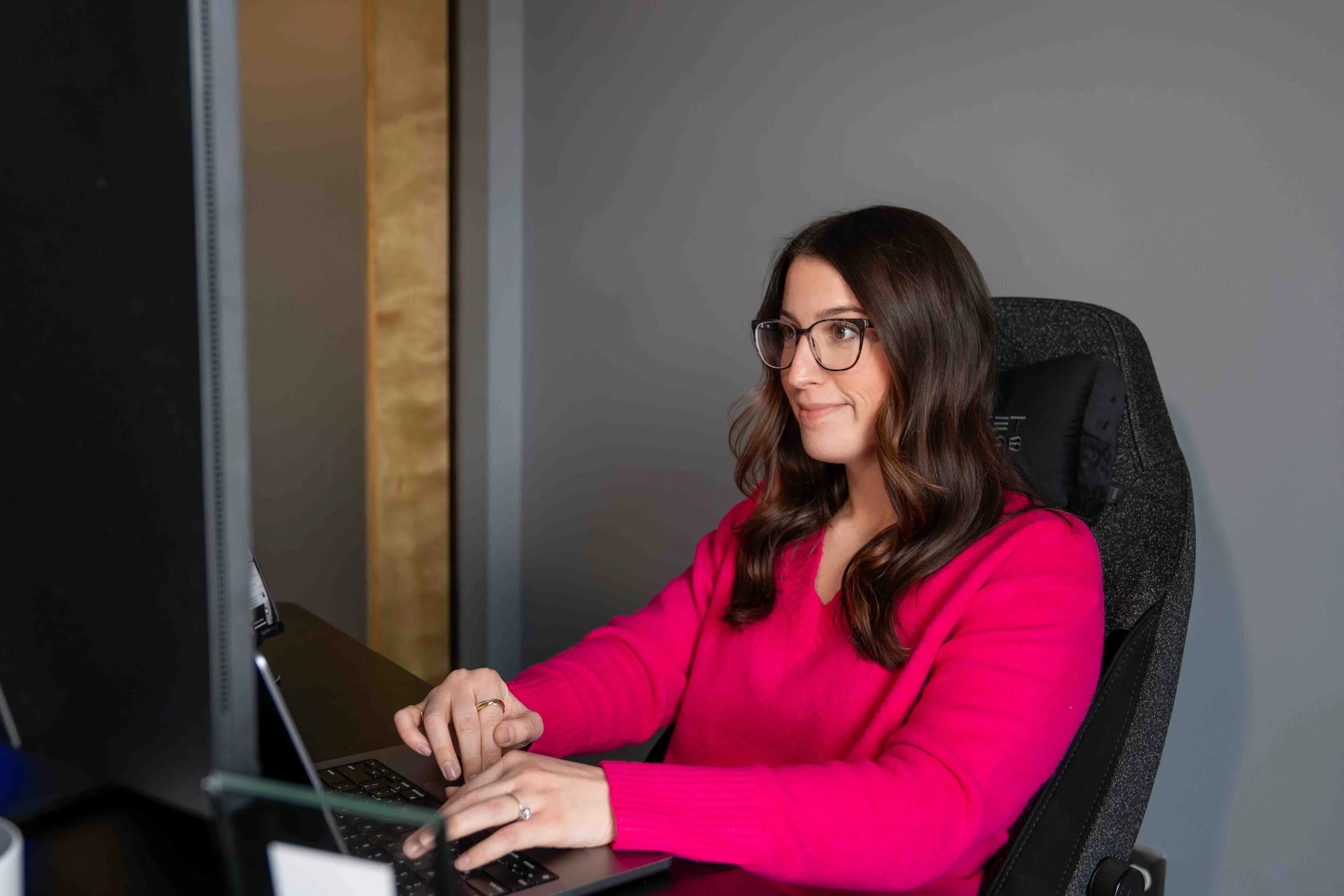 A woman with long brown hair and glasses working on a laptop at a desk.