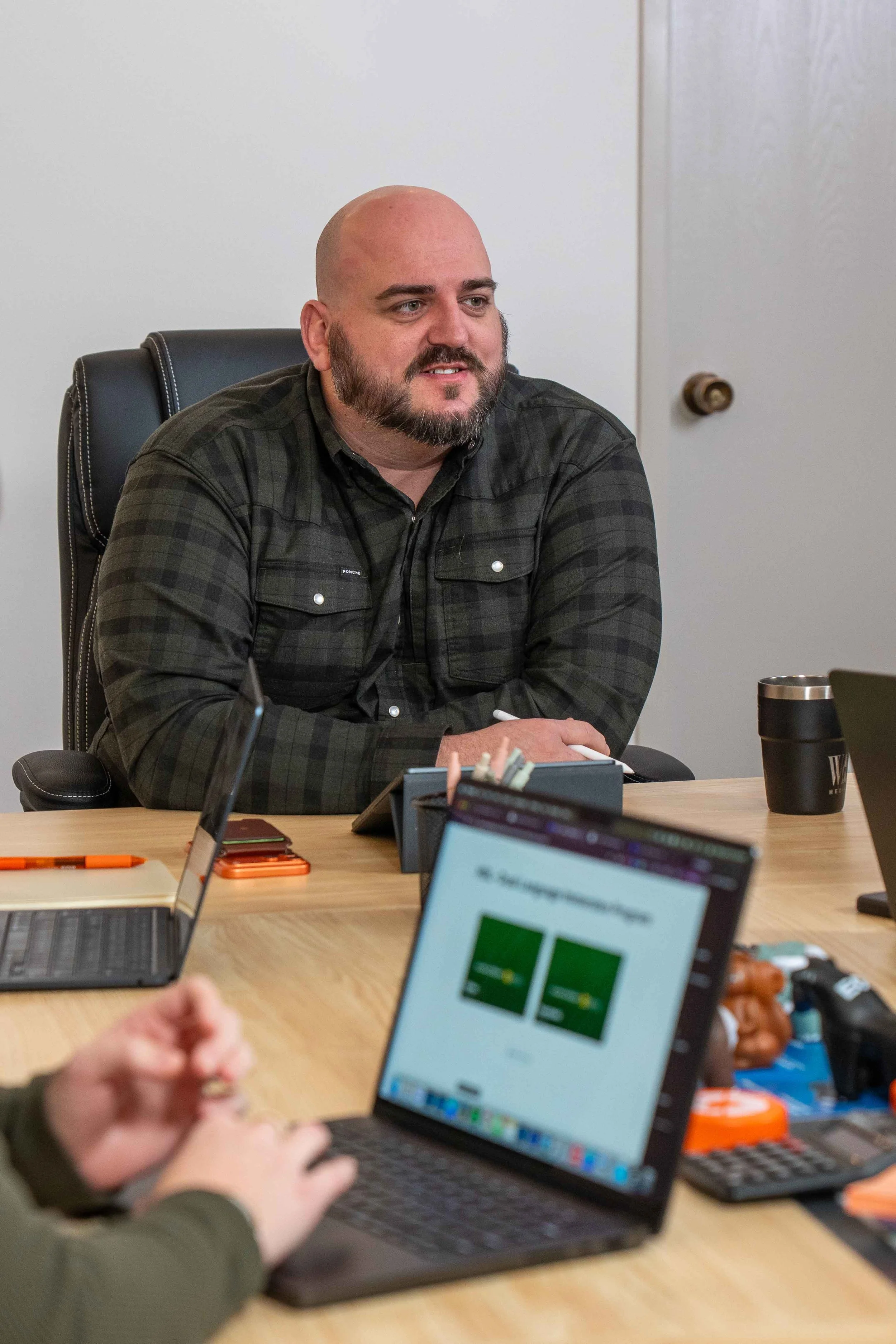 A man with a beard and a bald head sitting at a meeting table with laptops and office supplies.