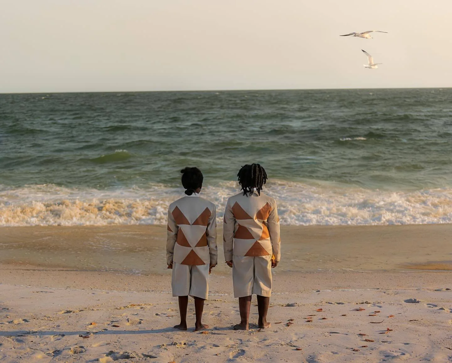 Malick Welli Two children in geometric-patterned clothing stand on a sandy beach facing the ocean, with seagulls flying above.