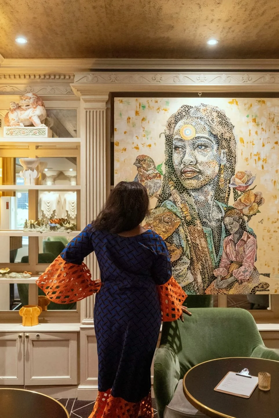 A woman viewing a large mosaic portrait of a woman with a bird on her shoulder in a well-decorated room with shelves of decorative items and a green armchair.