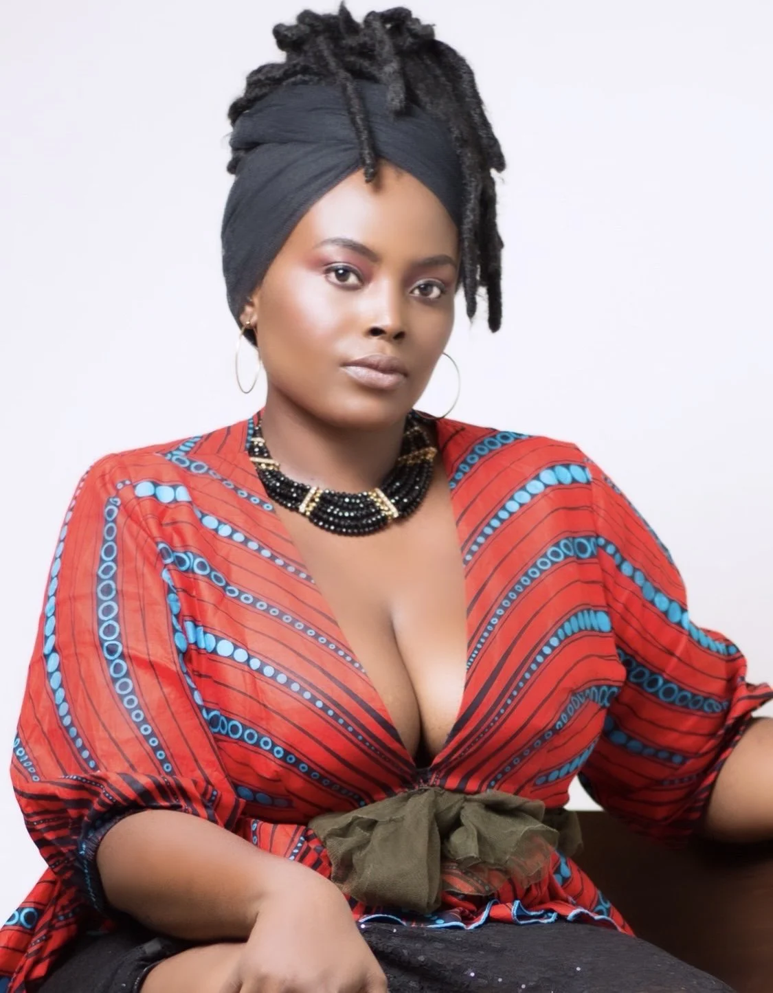 Portrait of a woman with dark skin, wearing a black headwrap with dreadlocks, a red patterned top, and gold hoop earrings.