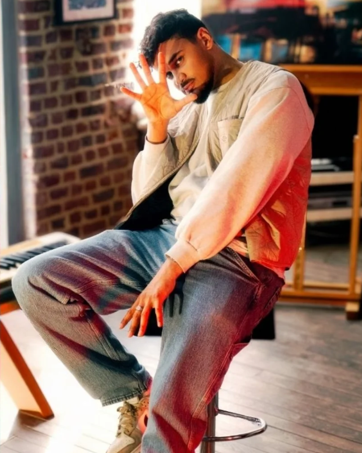 A young man with dark hair and a beard sitting on a stool in a room with a brick wall, bookshelves, and a keyboard, making a playful gesture with his right hand near his face and looking at the camera.